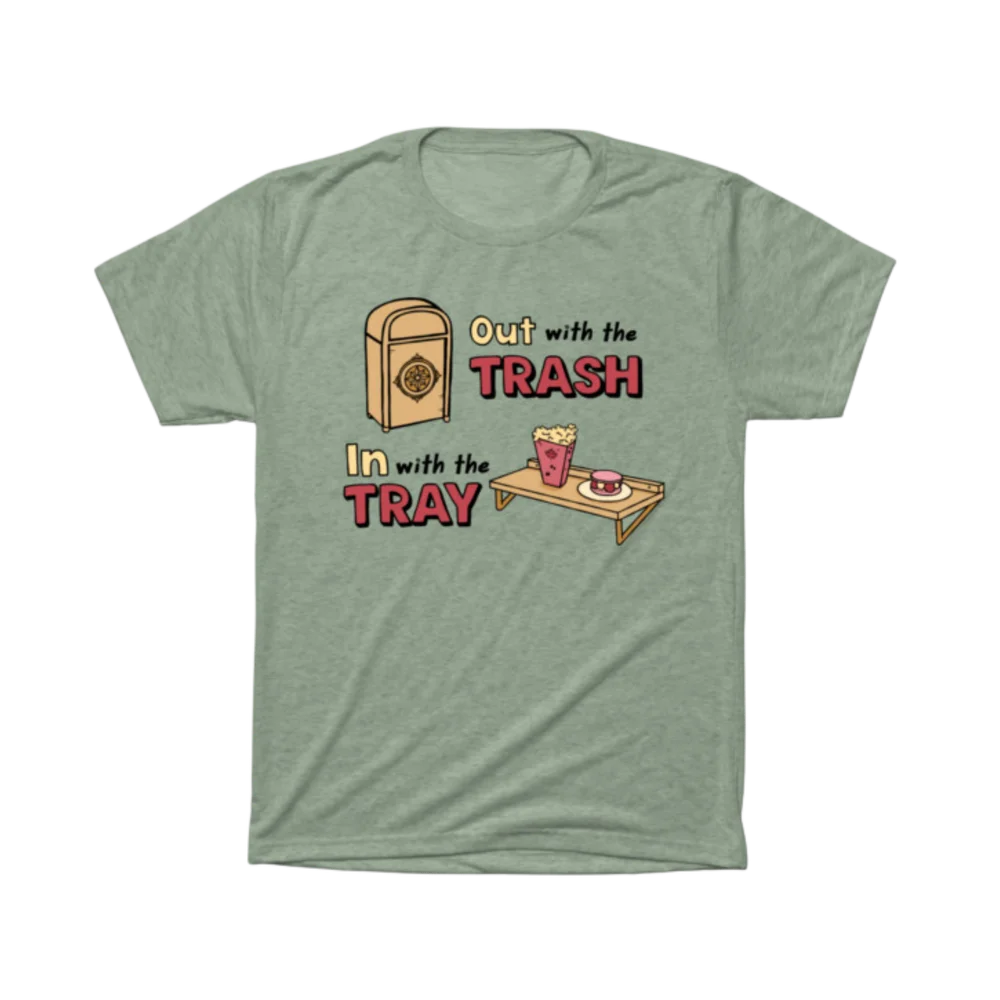 Sage green t-shirt printed with the Out With the Trash, In With the Tray design — a theme-park trash can next to a snack tray holding popcorn and a treat, playful lettering in cream and red.