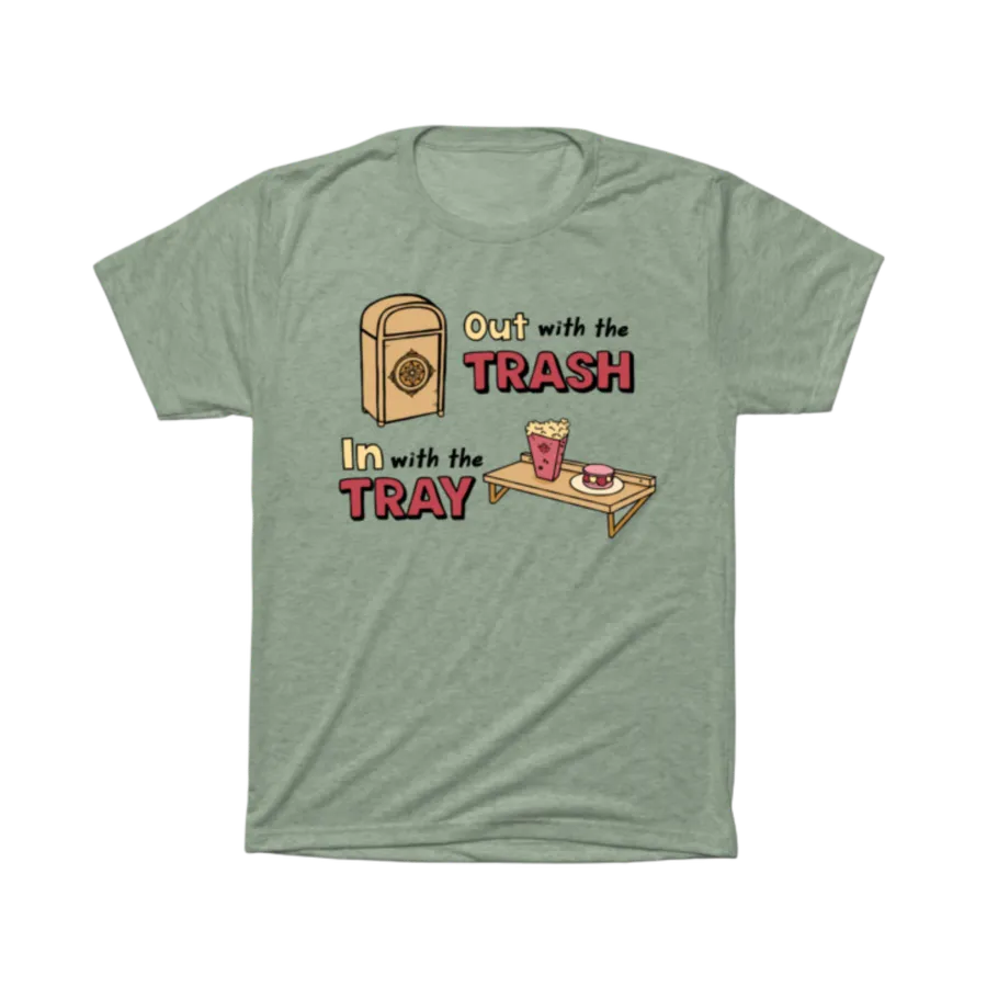 Sage green t-shirt printed with the Out With the Trash, In With the Tray design — a theme-park trash can next to a snack tray holding popcorn and a treat, playful lettering in cream and red.